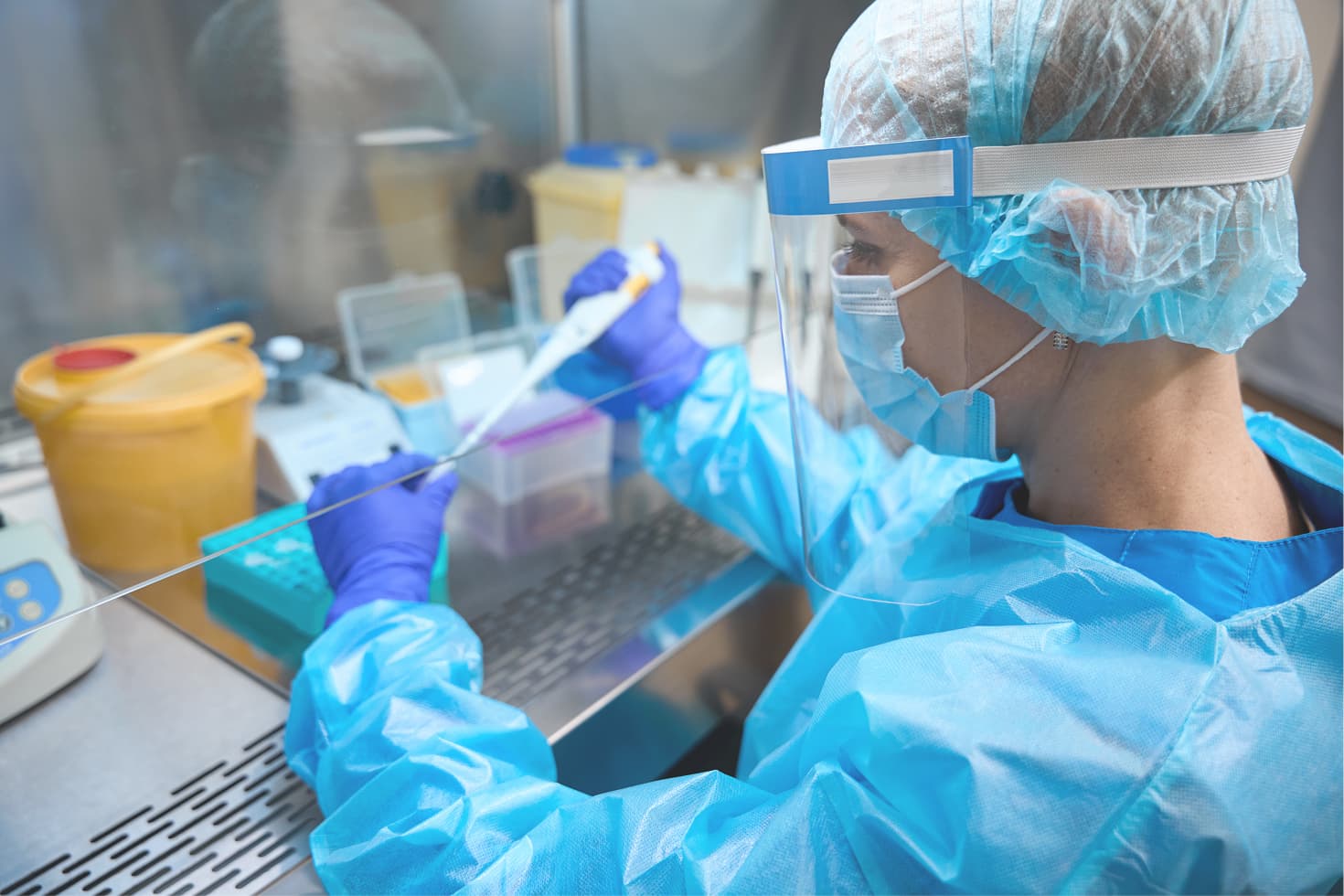 A scientist in protective gear works in a lab, using a pipette at a sterile workstation surrounded by lab equipment.