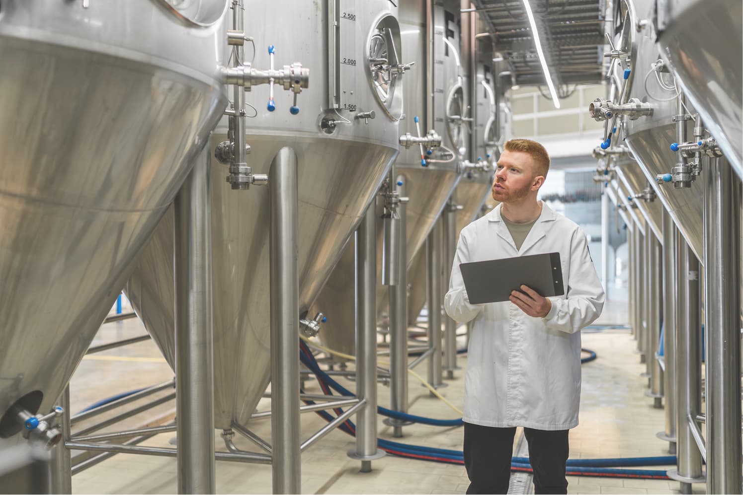A person in a white lab coat inspects large stainless steel fermentation tanks in a brewery or industrial facility, holding a clipboard.