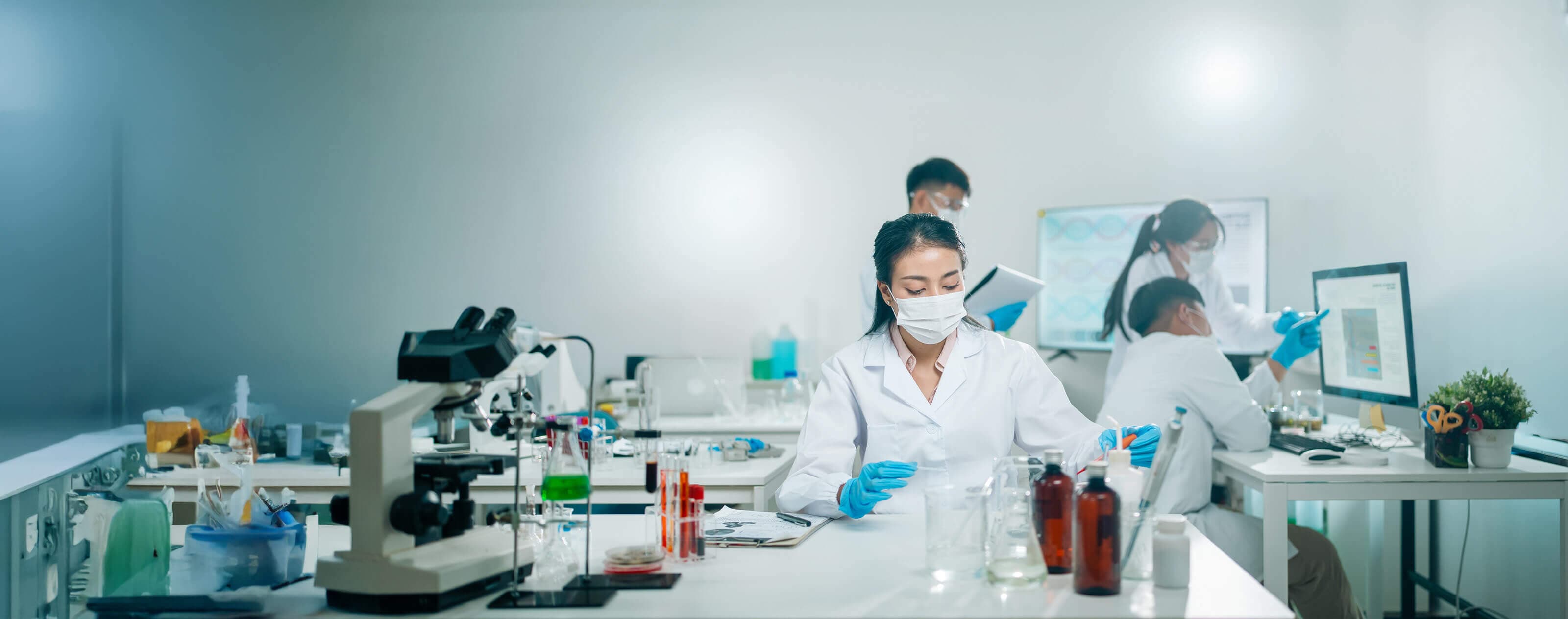 Scientists in a lab wearing masks and gloves conduct experiments, with microscopes and test tubes on the tables, and a computer in the background.