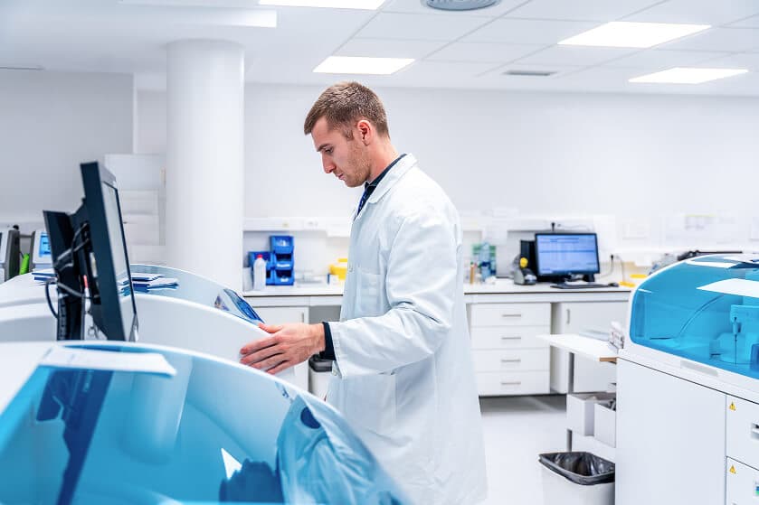 A scientist in a lab coat operates advanced laboratory equipment in a bright, modern lab with computers and various facilities in the background.